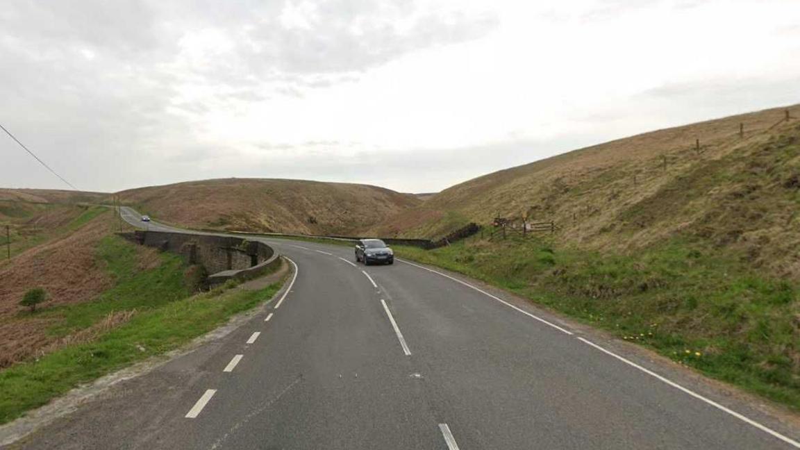 A road with grass verges either side and a car coming in the opposite direction. The road goes round a small corner.
