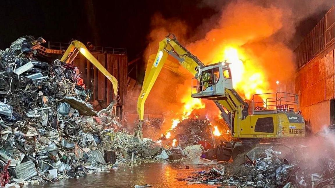 A fire burns within a scrap metal section of a recycling centre. A large pile of scrap metal sits to the left of the image. To the right is a large piece of machinery and the fire burns behind it. 