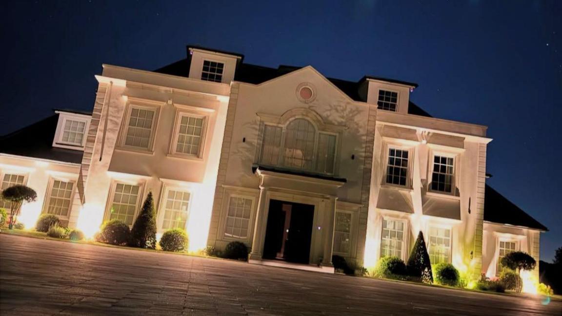 A large white house with bushes and a tiled driveway out the front. It is illuminated with lighting at night. There are lots of windows looking out towards the driveway, where the pictured was taken.