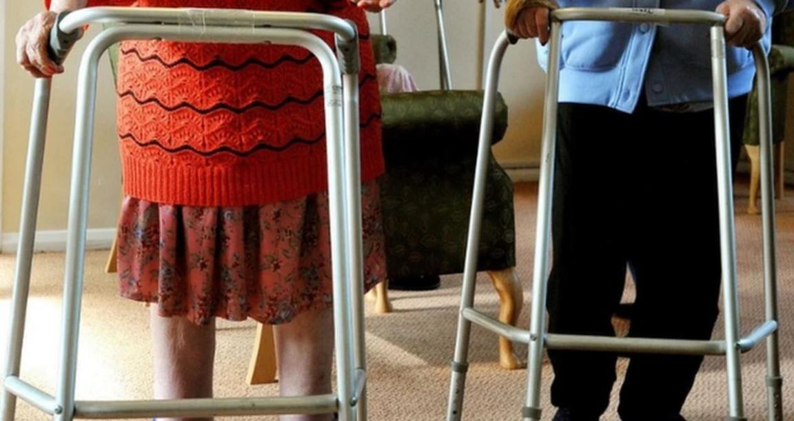 Two pairs of legs are seen behind steel walkers, one pair wears a red flowery skirt, the other black trousers. 