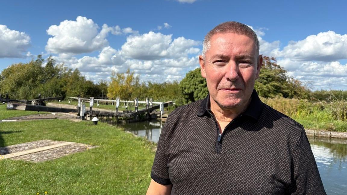 Stephen Golder with short dark hair looking at the camera and wearing a dark-coloured T-shirt. He is standing on a towpath with a canal and a lock behind him. There are trees and hedges in the background.