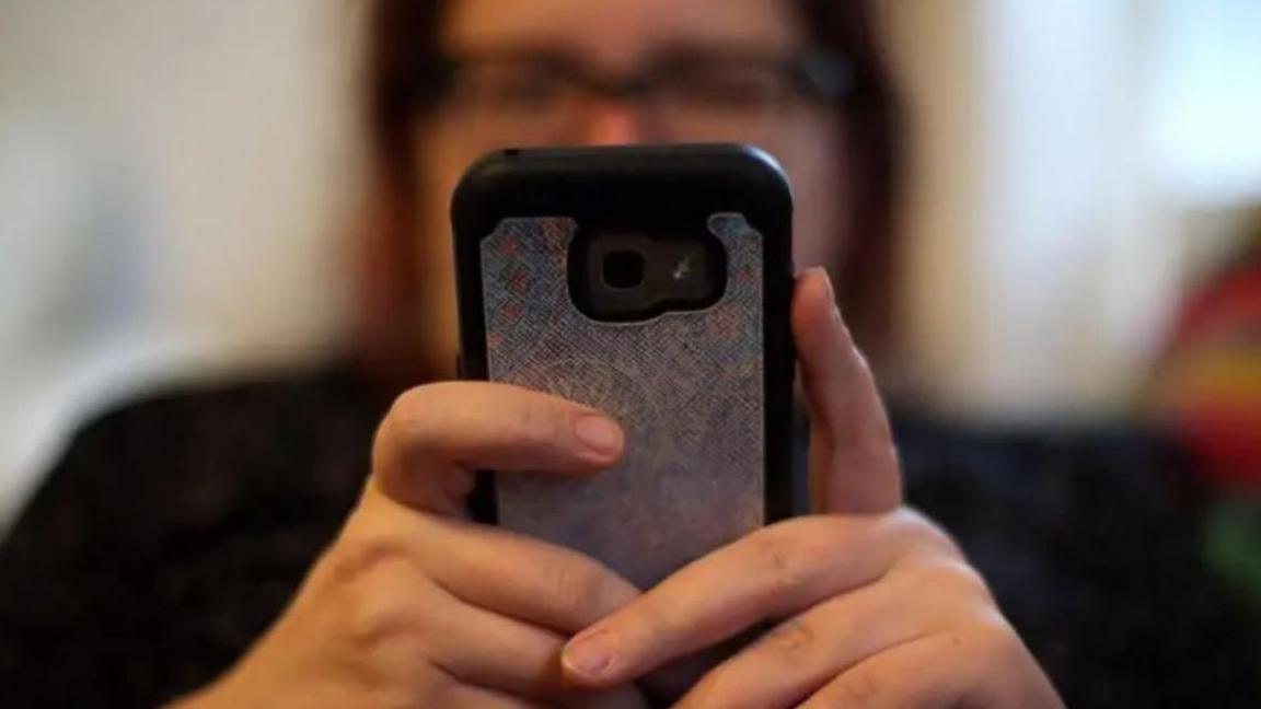 Woman blurred out wearing glasses holding a black mobile phone with grey sticker on the back in both her hands 