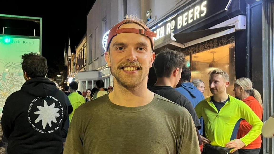 A man wearing a green T-shirt and a pink cap, which he is wearing backwards. He stands outside a bar amongst other runners at night time. He is smiling. 