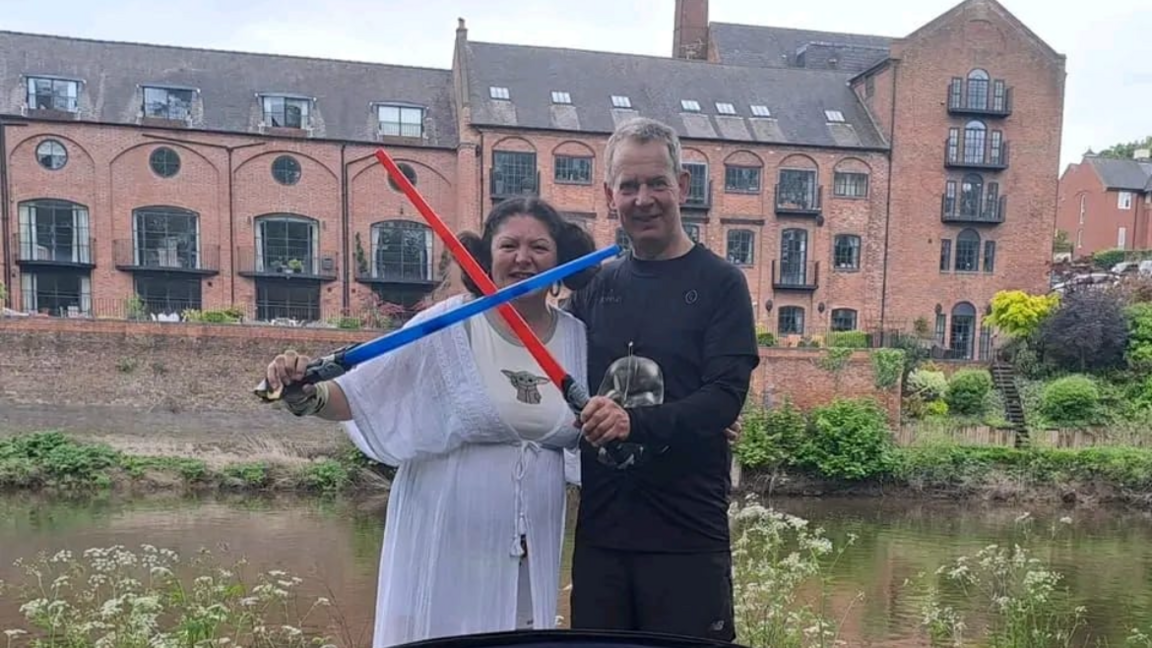 A woman dressed as Princess Leia in a white flowing top, with her hair in two big buns at the side of her head, holding a blue toy lightsaber, which is crossed with a red lightsaber held by a man next to her in black clothing. They stand in front of the river and a row of red brick buildings