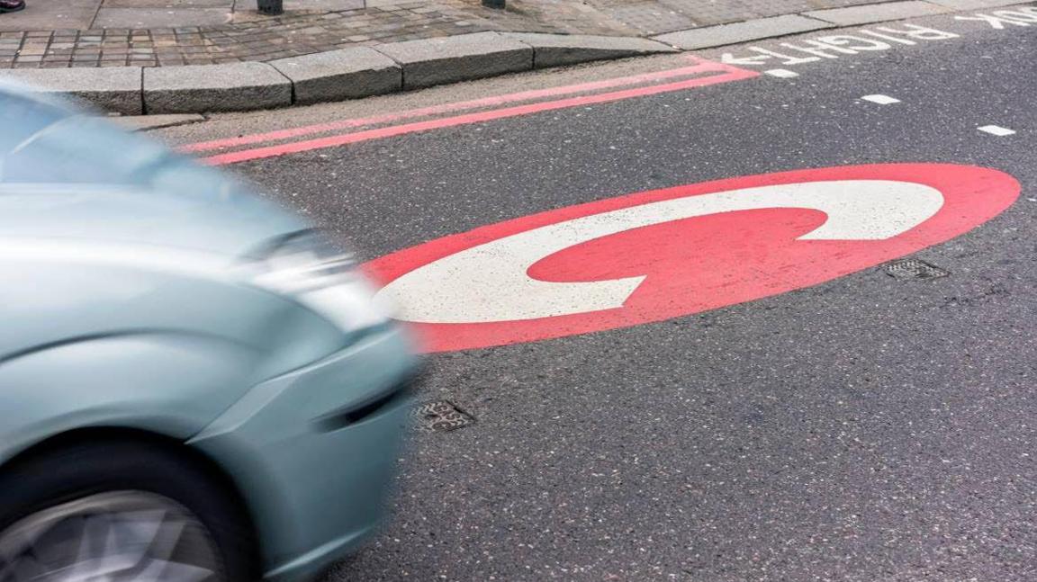 A red circle with a large white letter C is painted on the surface of a road in front of a crossing. A silver-blue car is driving towards the painted C from the left, and is blurred from motion. The pavement with double red lines along the kerb is visible in the background.
