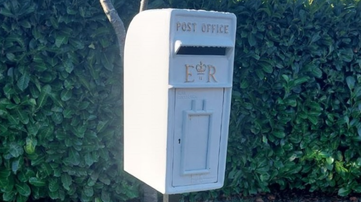 A postbox painted white with gold lettering. Behind it is a hedge.