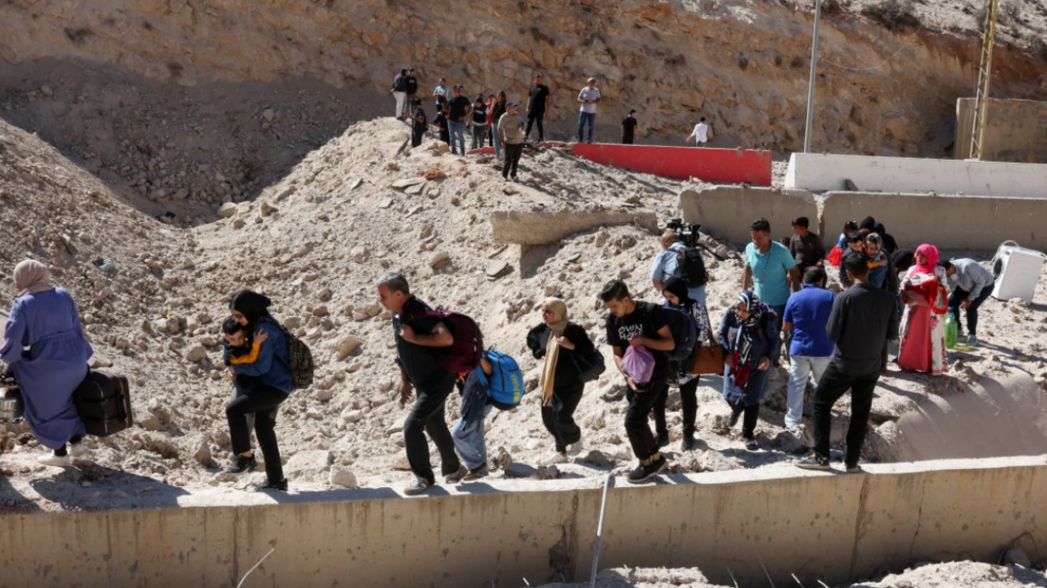 Dozens of people carrying their childen and belongings walk through the cratered road to reach the Masnaa crossing taking them into Syria on 4 October 2024.