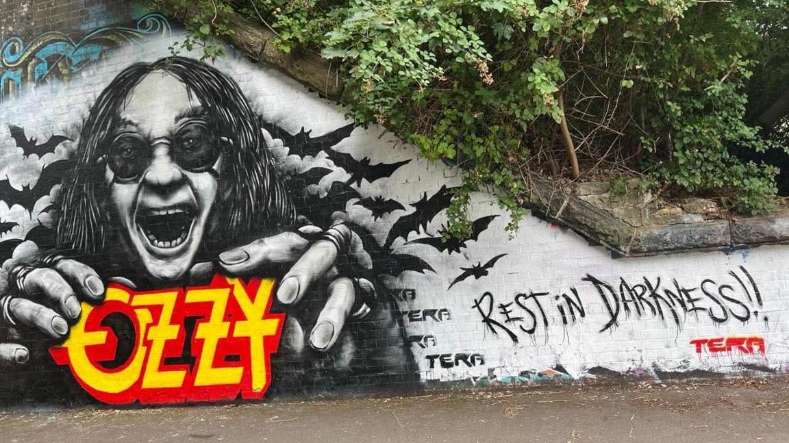 A large colourful mural of Ozzy Osbourne is seen in St Werburgh's in Bristol. He is in his familiar pose with his mouth wide open and hands reaching out. Behind him are drawn some black bats, and he is wearing dark glasses. "Rest in Darkness" has been written next to him in black