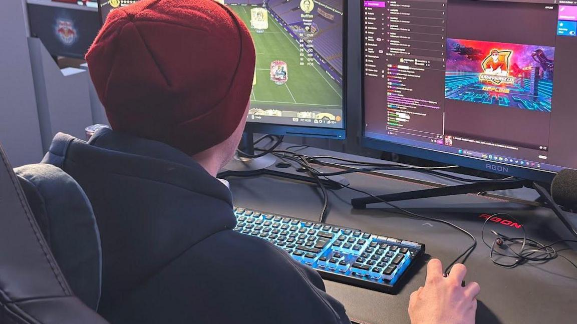 A man faces away from the camera with his hands on a keyboard and mouse facing two computer screens. He's wearing a red beanie hat