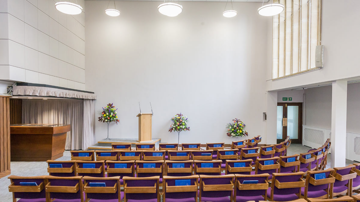 The interior of Worcester Crematorium's chapel. Seats are arranged in front of a lectern and the room is lit by lights hanging from the ceiling.