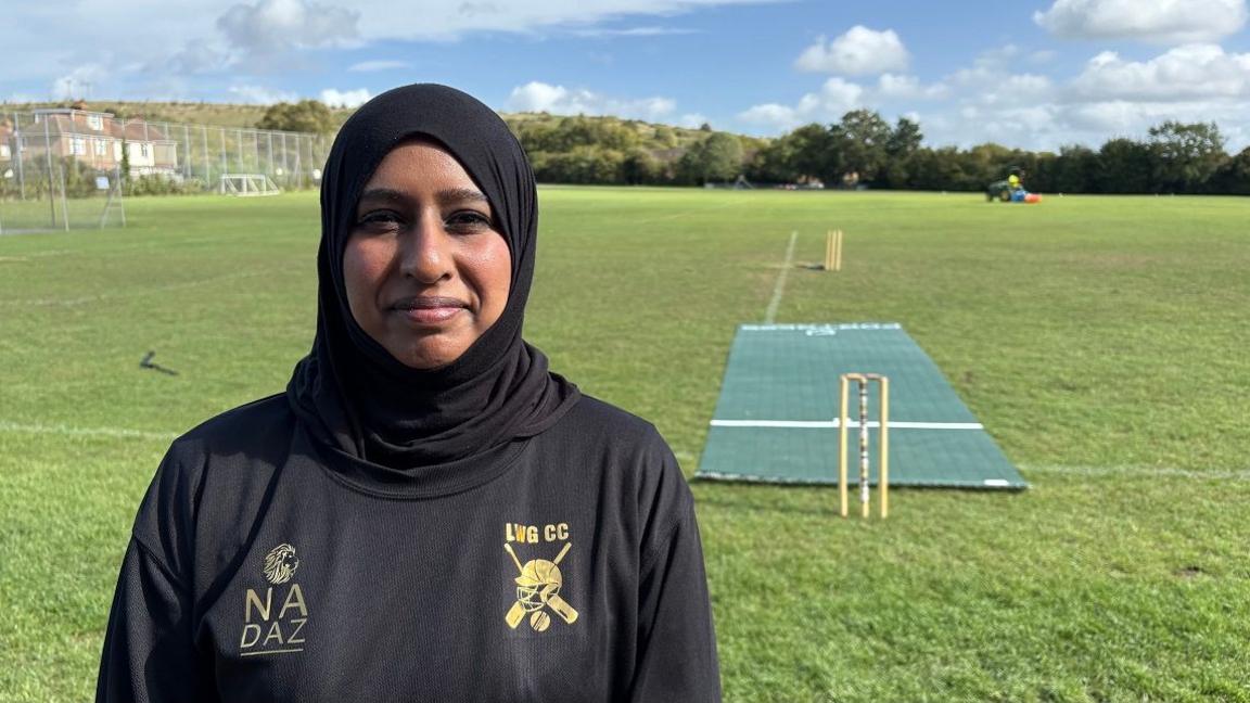 Mahira Hussain with a black hijab and black top with LWGCC logo and Nadaz logo. She is standing on a cricket pitch with wickets and a fabric cricket square behind. There is a mower in the distance, and hedges beyond.