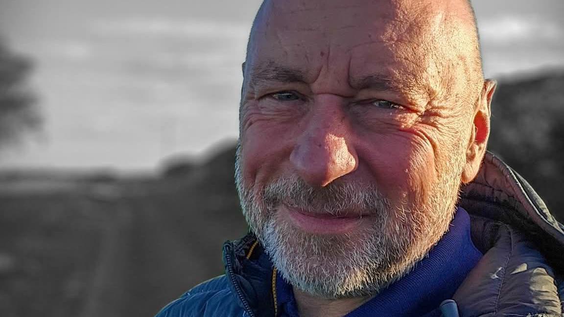 Billy Millward, aged 55, looks at the camera. He is standing in front of a country lane which has been shaded black and white in the background. He is bald with greying stubble on his beard and upper lip. He is wearing a grey anorak with a blue fleece underneath. The sun is shining on the left of his face. The right of his face is shaded.