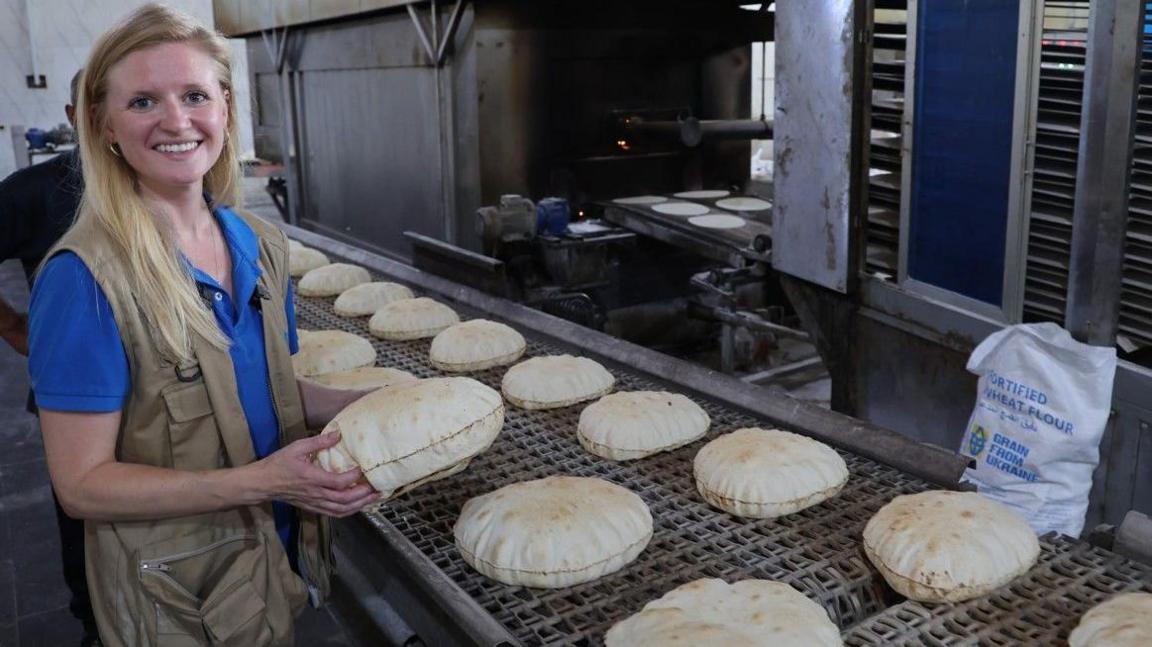 A young caucasian woman with long blonde hair wears a bright blue World Food Programme Tshirt and smiles at the camera in front of a conveyorbelt full of freshly baked round bread