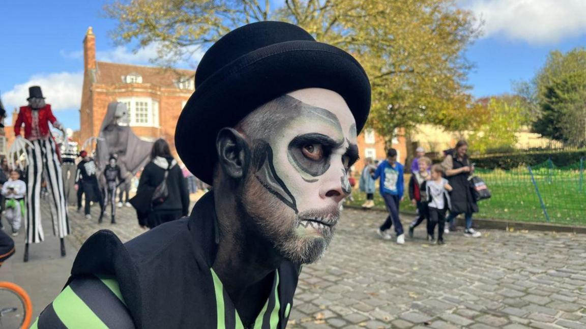 A man with skeleton face paint on and a green and black skeleton outfit side eye'ing the camera in the street. 