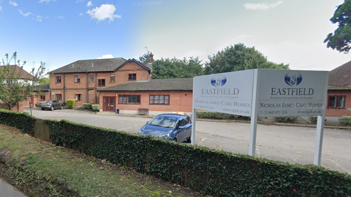 A red brick building and car park sit behind a green hedge. There is a parked car and a sign which says "Eastfield" in the foreground. 