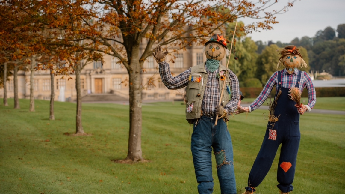 Two scarecrows in front of Longleat House in checked shirts and patched jeans with smiles and one waving to camera 