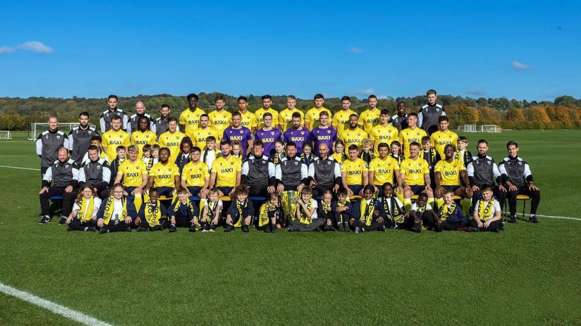 A football quad wearing a mix of yellow, purple and black kits stand in three rows on a football pitch to have their picture taken. A row of primary school children sit on the grass in front wearing yellow Oxford United scarves. It's a sunny say and the sky is blue.