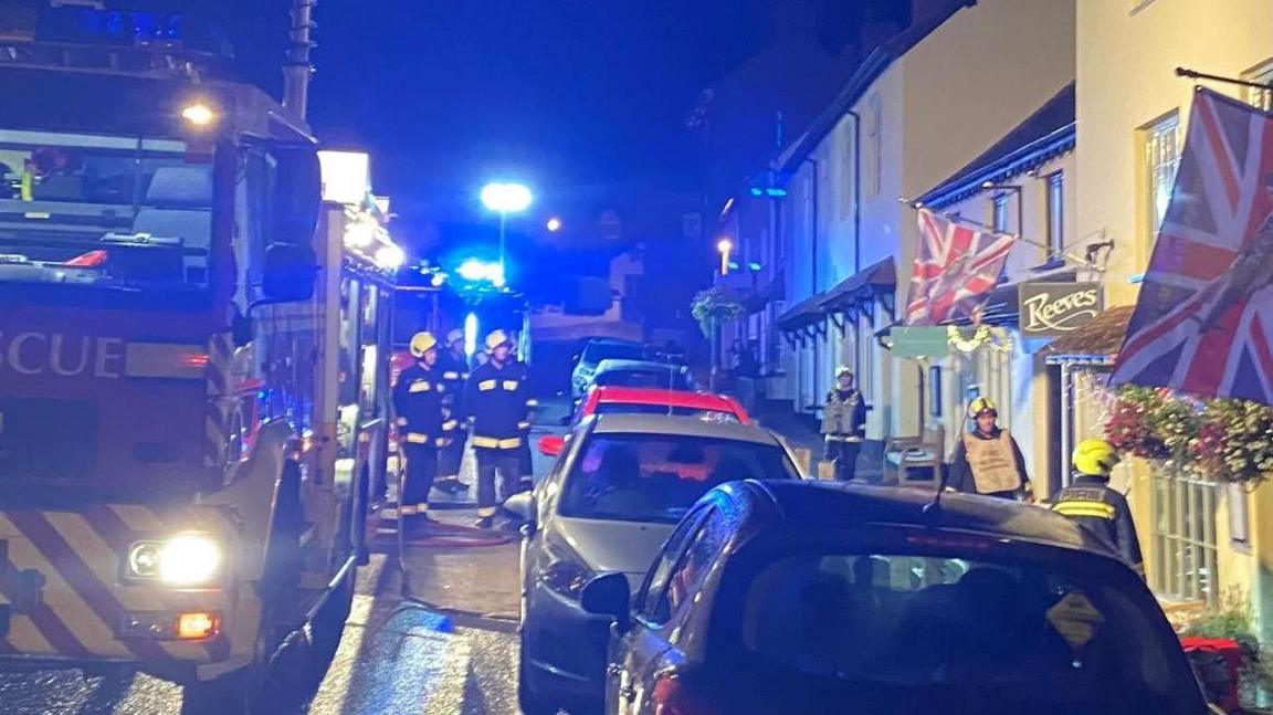 The outside of the Reeves restaurant with a fire engine parked on the street. Fire fighters in uniform are standing next to the engine. It is night time.