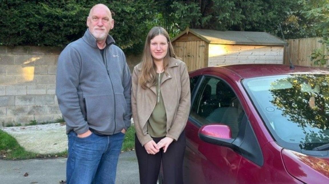 Carl Goodwin on the left and Zoe Johnston standing next to a car, with a shed in the background, in front of a wall that has a line of trees above it.