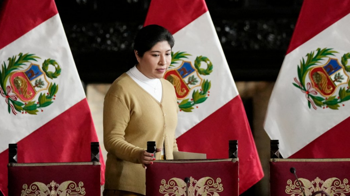 Betssy Chavez wearing a white turtleneck shirt and a buttoned-up, beige cardigan. Her right hand is gripping the top of a wooden chair, three Peruvian flags attached to poles behind her.