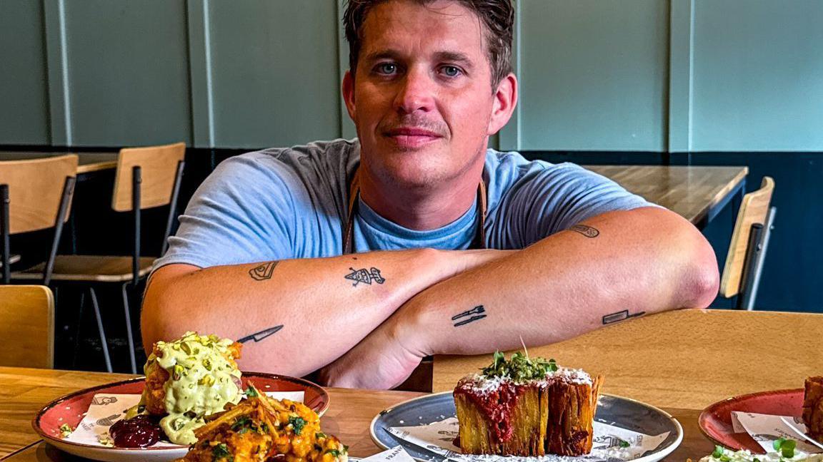 Adam crouches to table level with his arms resting on the back of a wooden chair. On a wooden table in front of him, three plates of food are placed. Behind him, other wooden chairs and tables are set out in the room. 