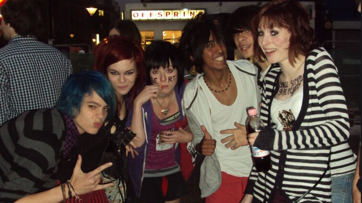 Six people take a picture outside an Offspring concert. The person on the left has blue hair and a black and grey striped cardigan. The girl to the right of him has big red hair and black eyeliner. A girl in the middle has a straight black fringe and wears a purple hoodie and pink top. She has writing on her face. To her right a man with a straight black fringe, grey hoodie and white vest smiles at the camera. In the front right a girl has wears a black and white cardigan, she has writing on her chest and carries a bottle. She has short brown hair and black eyeliner.