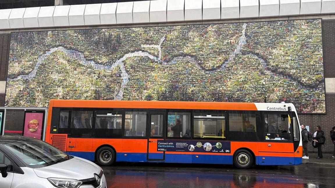 An exterior wall of a shopping centre showing a montage of thousands of pictures of people arranged around a river running across the centre of the picture. There is a single-decker red and blue bus in front of the montage and people are queueing to get on the bus.