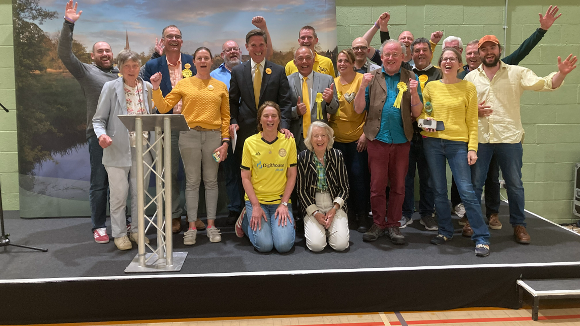 Charlie Maynard - wearing a suit with a yellow tie and yellow Lib Dem sticker - celebrates on the podium where results were announced in Witney 