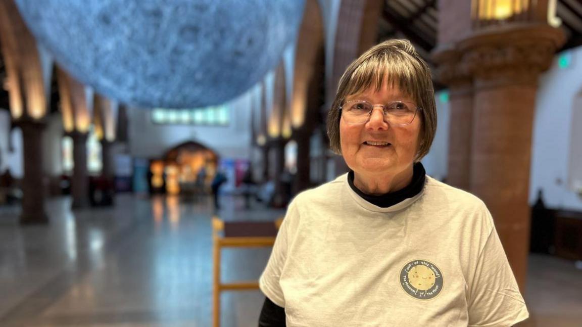 Rosemary Clarke smiling. She has short grey hair and is wearing glasses and a wearing a white T-shirt. The sculpture of the moon is blurred out in the background.