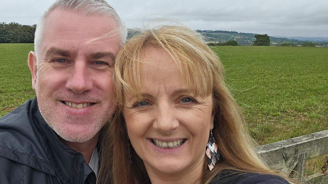 Justin (left) with grey hair and stubble wears a blue jacket hugs Gail Hardy (right) who has long blond hair and dangly bronze and silver leaf style earrings. They are standing in front of a field and are both smiling.