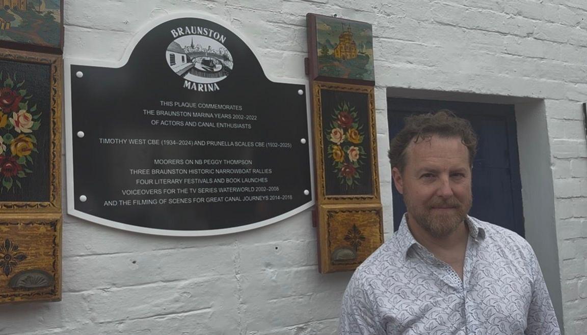 An image of actor Samuel West standing next to a black and white commemorative plaque at Braunston Marina, surrounded by traditional "Roses and Castles" style painted canalware panels.