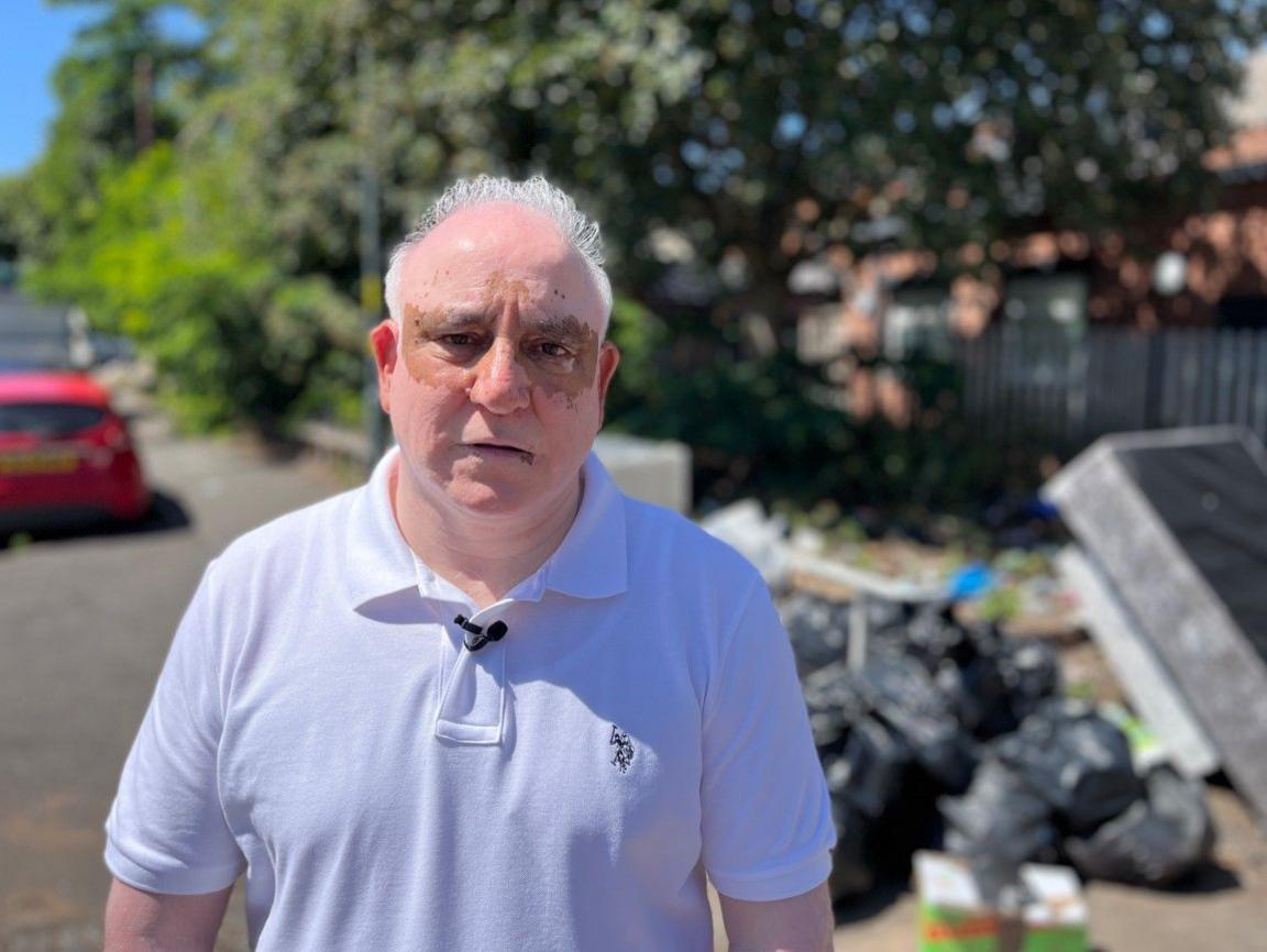 A man with white hair and a white top is looking at the camera. Black bin bags are blurred on the photo on the pavement on the right.