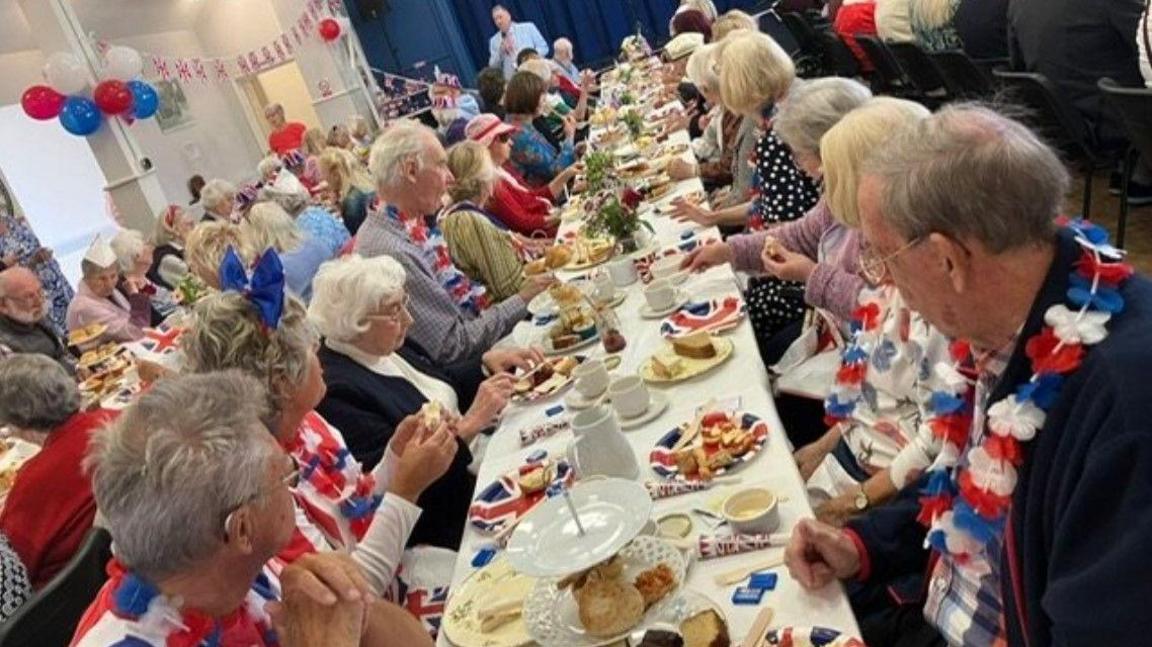 Scores of people sitting at long tables filled with plates and food. Many are wearing red, white and blue garlands around their necks and the paper plates are printed with the union flag.
