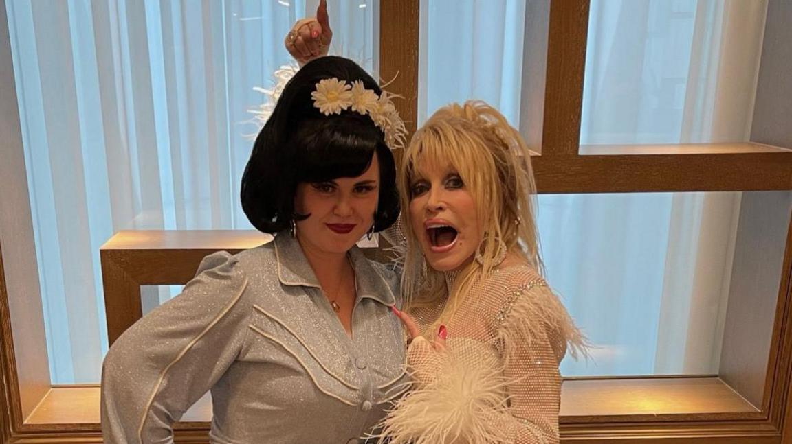 A photo of the top half of Sarah Wood - on the left - and Dolly Parton. Sarah has black hair, white flowers in her hair and is wearing something grey and glittery. Dolly Parton is pointing at Sarah, and is wearing a pale outfit with diamantes and feathers on. 