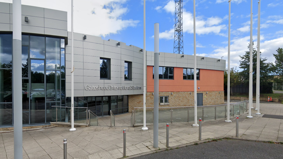 The entrance to Gateshead Stadium. It has grey cladding with the words "Gateshead International Stadium" on it and glass windows. The right-hand side of the building is made from brick.