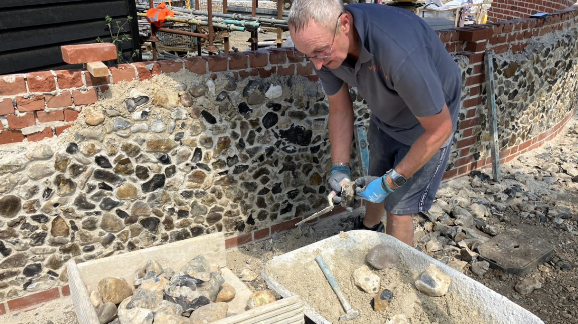 Mark Clifton, who has short grey hair and glasses and is wearing a grey polo shirt and blue work gloves, is standing next to partially constructed flint wall. He is bending at the waist and looking at a wooden box filled with flints, while holding one in his hands, along with a trowel.
