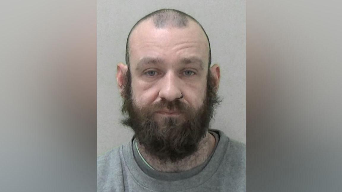 Mugshot of Daniel Devlin, he has a shaved head and a big but unkempt bushy beard, with a ring in one of his nostrils and is wearing a grey sweatshirt