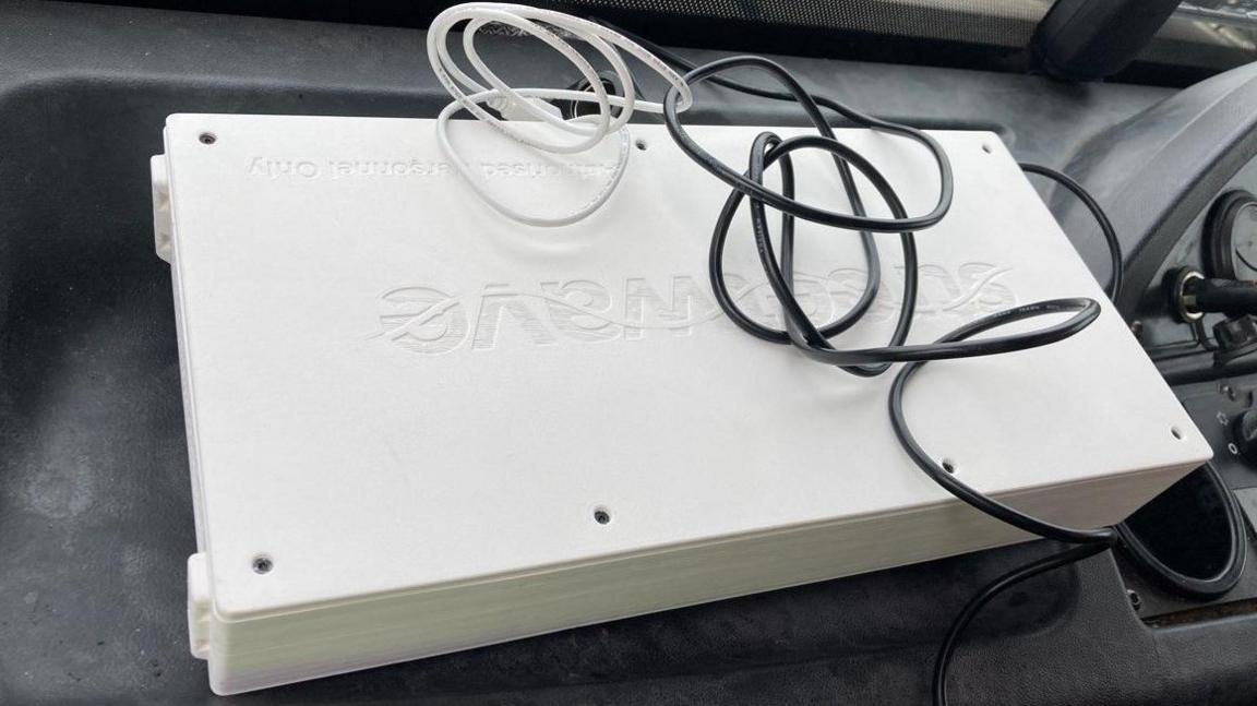 A white plastic box with black and white wires attached to it sits on the dashboard of a vehicle