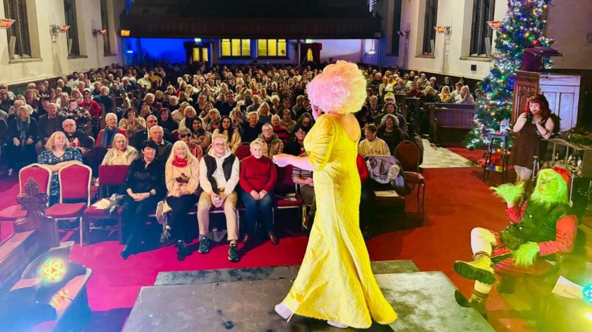 A drag queen in a large blonde wig wearing a full-length yellow ball gown is stood entertaining a seated audience. She is not facing the camera. A person dressed as the Grint is sat to the right of her.