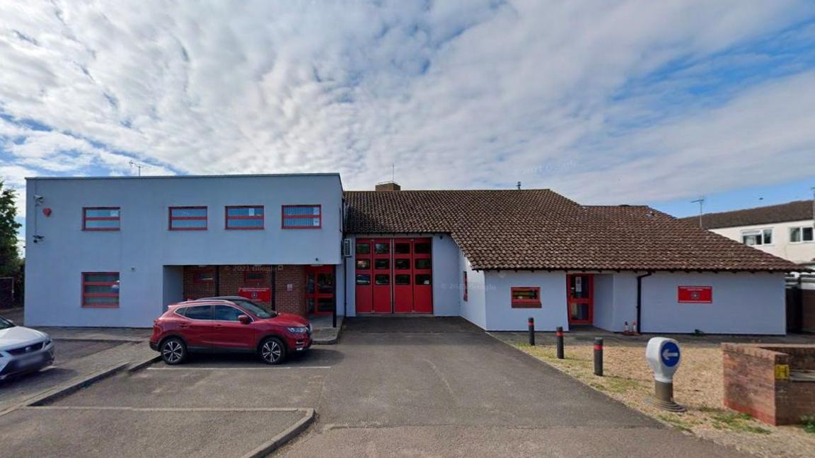 Two-storey fire station painted white with small red-framed windows. There are large red double doors in the centre of the building and red pedestrian access doors to the left. There is also a small red door in the single-storey section of the building to the right. There is a red car parked in front of the pedestrian access doors.