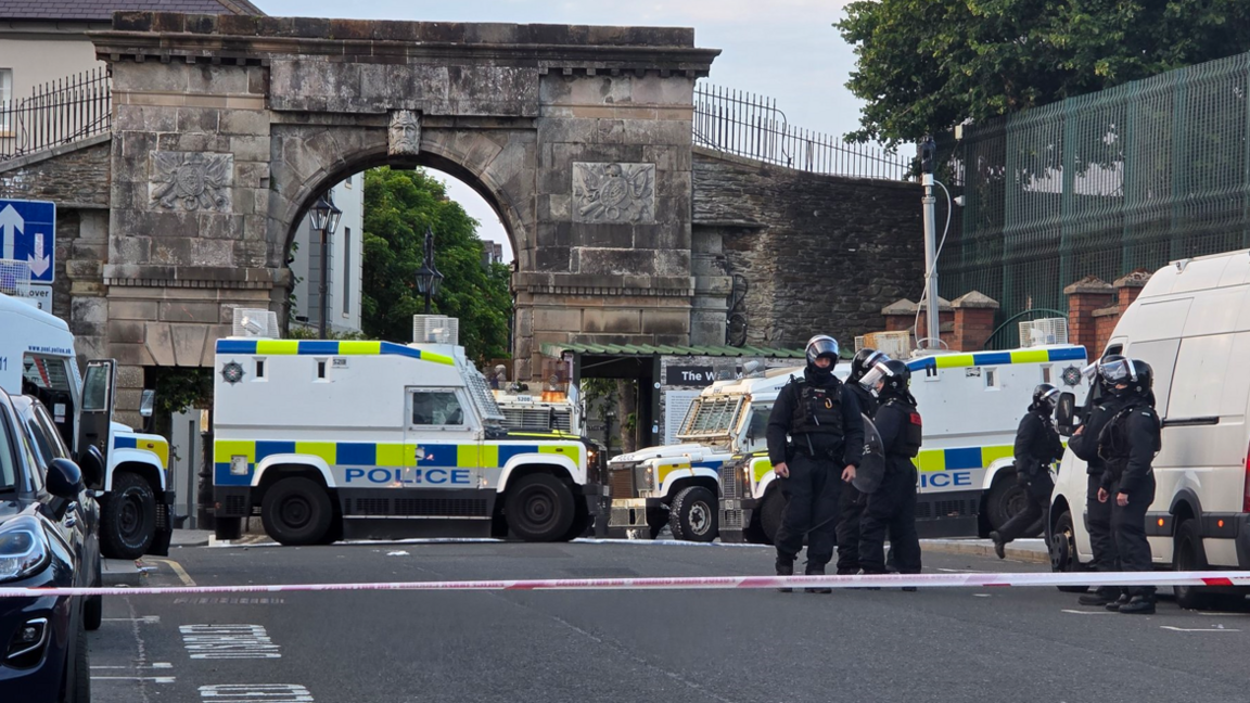 Shows several police Land Rovers and riot police in black and wearing helmets in front of a police tape and a historic arch on the city walls