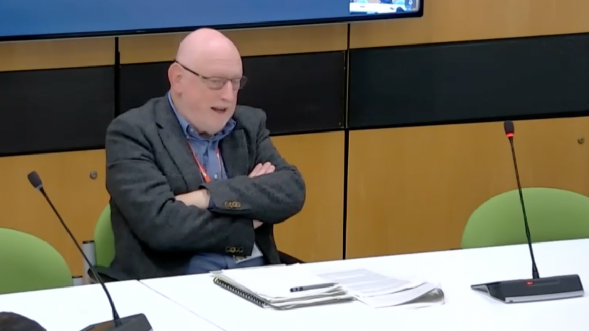 Lead Commissioner Max Caller sits with arms folded in front of notes and microphones at the meeting of the council's scrutiny committee.