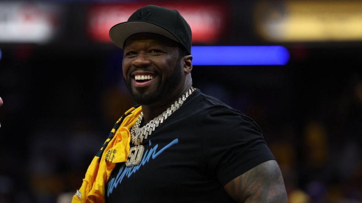 50 Cent is pictured smiling and looking off camera. He is wearing a black baseball hat, a black T-shirt and diamond-encrusted silver chain with the numbers "50" hanging down.