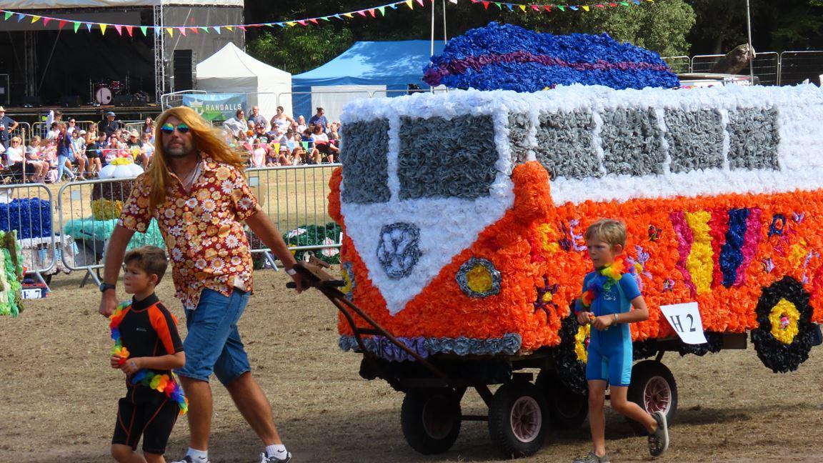 A man dressed in a flowery shirt, a long blonde wig and sunglasses pulls his VW Bus display along the showground at the North Show's Battle of Flowers in Guernsey. Two boys wearing wetsuits are walking next to the display, which mainly has orange and white flowers.