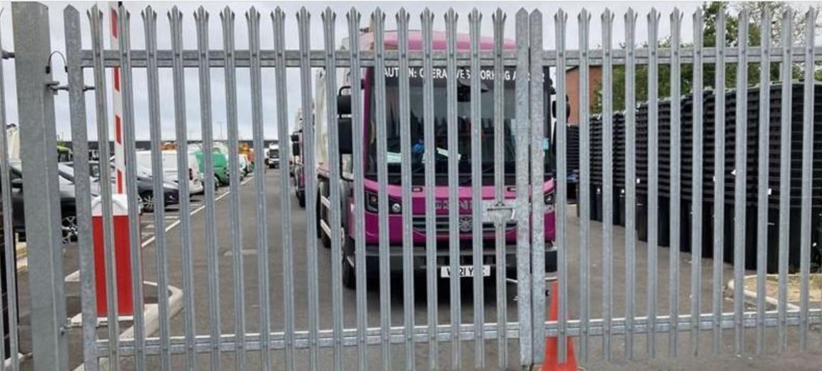 A purple waste lorry sits behind metal gates. An orange cone can be seen in front.