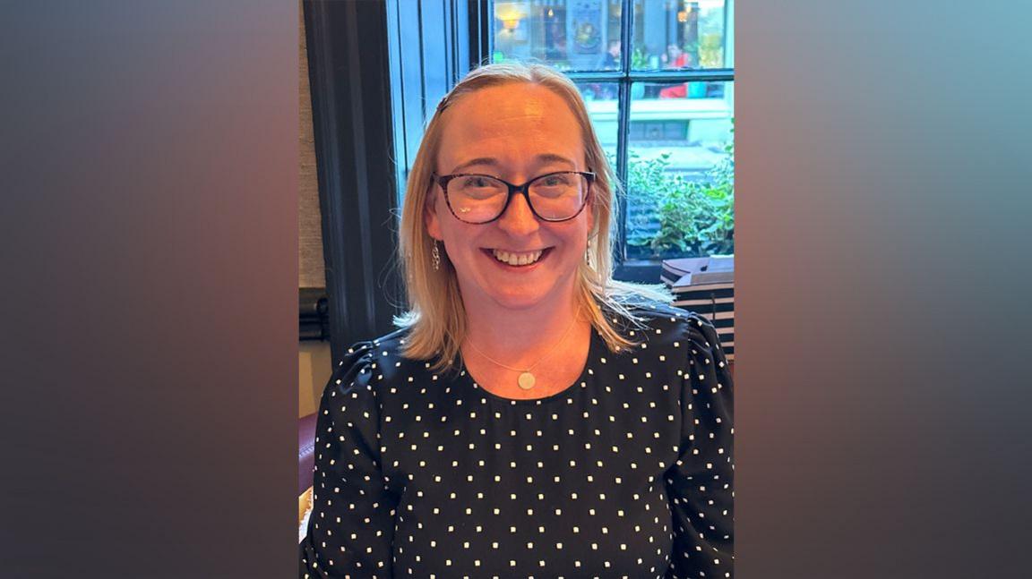 Nicola Packer wearing a spotty top smiles at camera. She is blonde with glasses.