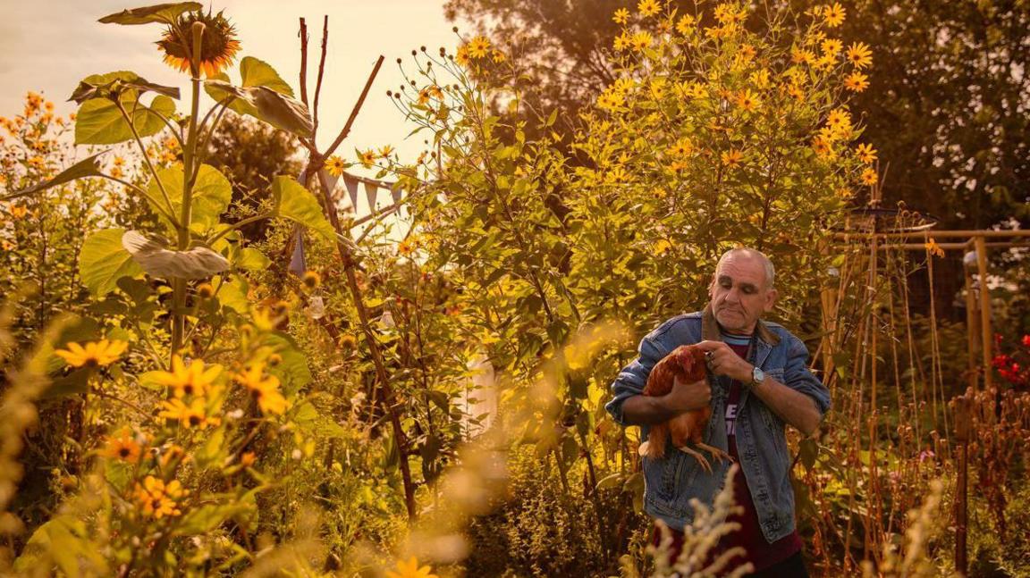 Photo of a man holding a large bird in a wood surrounded by flowers