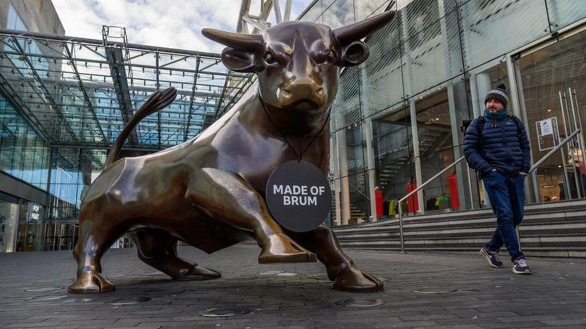 The Bullring bull is prominent on the left and in the middle of the photo. A man in blue on the right is looking at it.
