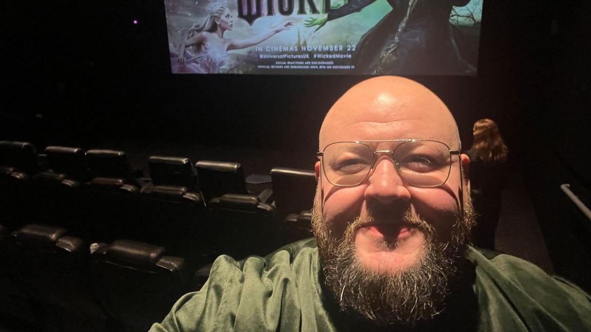 A man with bald head and a beard, wearing a green top. He is stood in a cinema with a large screen and rows of seats behind him. There is a still on the screen that says 'Wicked in cinemas November 22'.