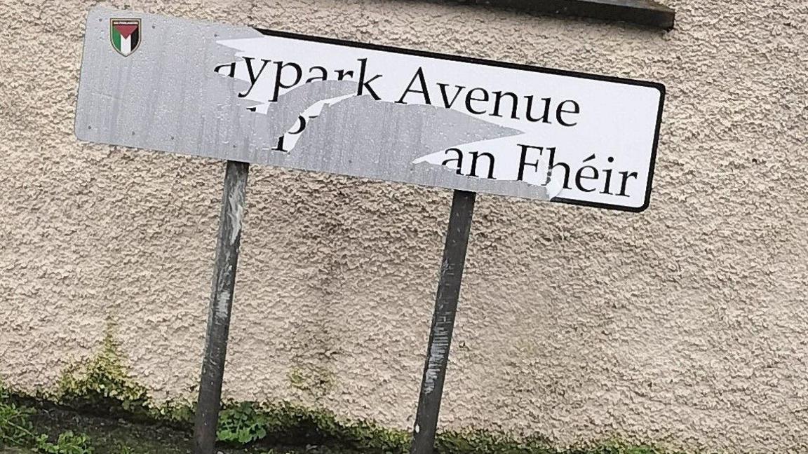 A damaged black and white dual-language street sign in south Belfast which is positioned in front of a cream wall.